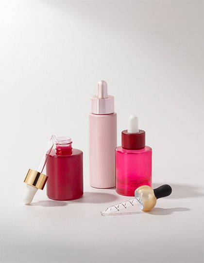 Collection of pink and reddish cosmetic containers with various droppers in different finishes—matte, glossy, gold, and colored—displayed on a grey background