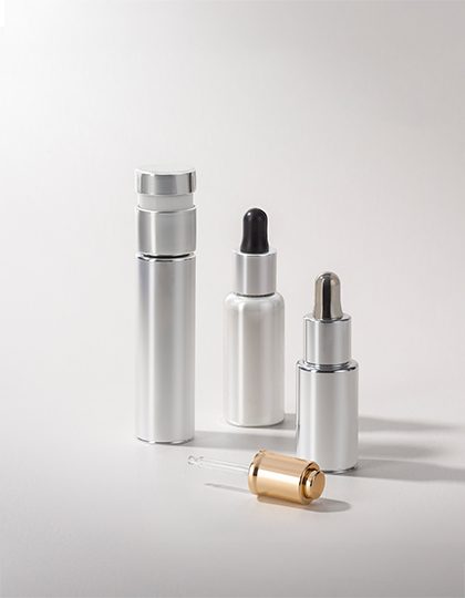 A collection of silver and gold cosmetic containers with various dispensing mechanisms, including droppers and pumps, displayed on a neutral background.