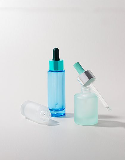 Collection of molded glass containers in various sizes, shapes, and finishes—some matte, others transparent, and some glossy—with different push-button dispensing mechanisms, displayed on a grey background.