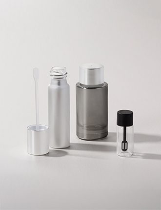 Collection of glass containers in various sizes with metallic and transparent finishes, accompanied by spatulas and caps, displayed on a neutral gray background.