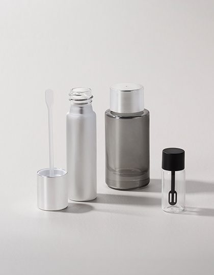 Collection of glass containers in various sizes with metallic and transparent finishes, accompanied by spatulas and caps, displayed on a neutral gray background.